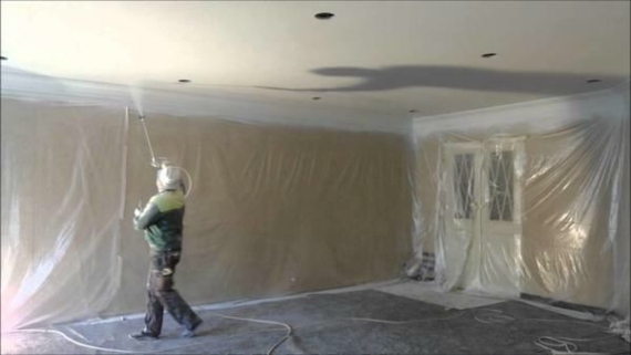 Homem pintando teto de uma residência com pistola de tinta spray usando todos os equipamentos necessários e com todo o restante do ambiente forrado 